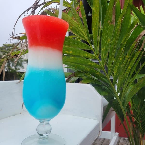 Red, white, and blue frozen drink in a tall glass on a white railing with palm leaves.