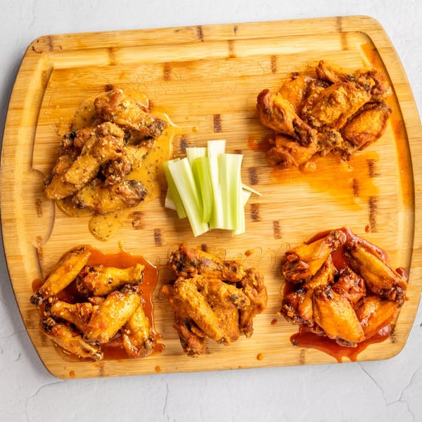 Five piles of House Of Sauce chicken wings with different sauces and celery sticks on a wooden board.