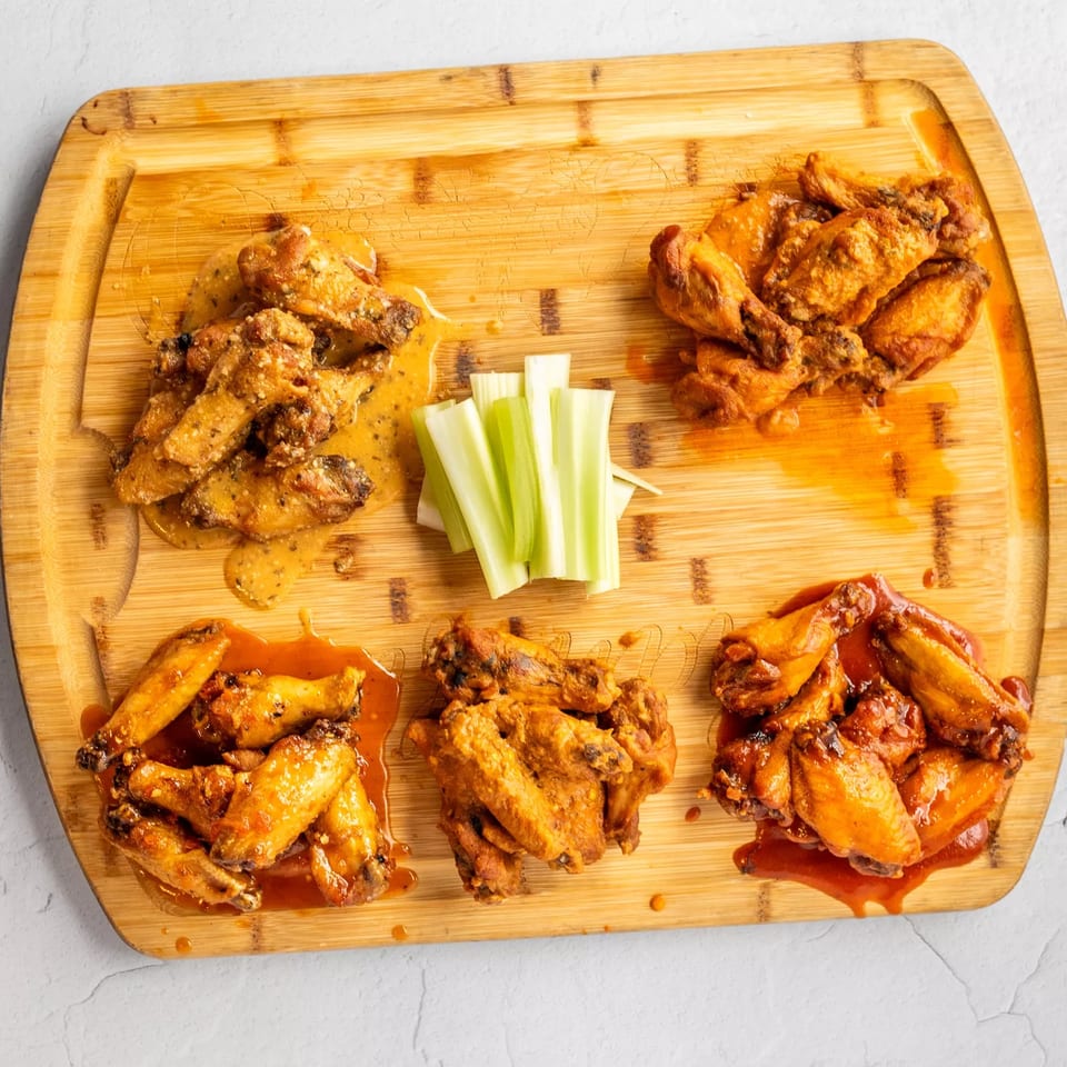 Five piles of House Of Sauce chicken wings with different sauces and celery sticks on a wooden board.