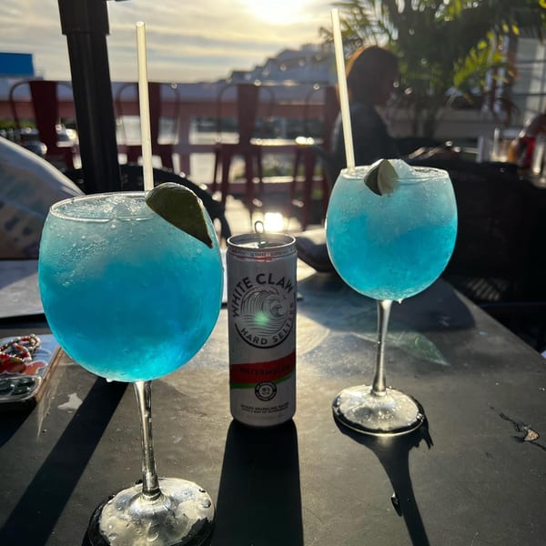 Two blue cocktails with lime and a White Claw seltzer on an outdoor patio at sunset.